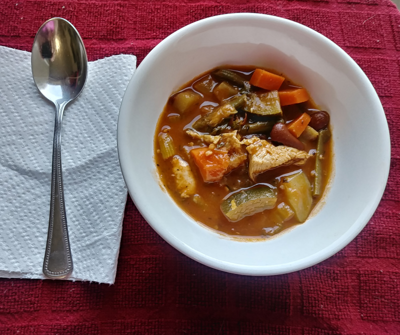 Chicken minestrone in a white bowl on top of a red place setting with a spoon sitting next to it. 