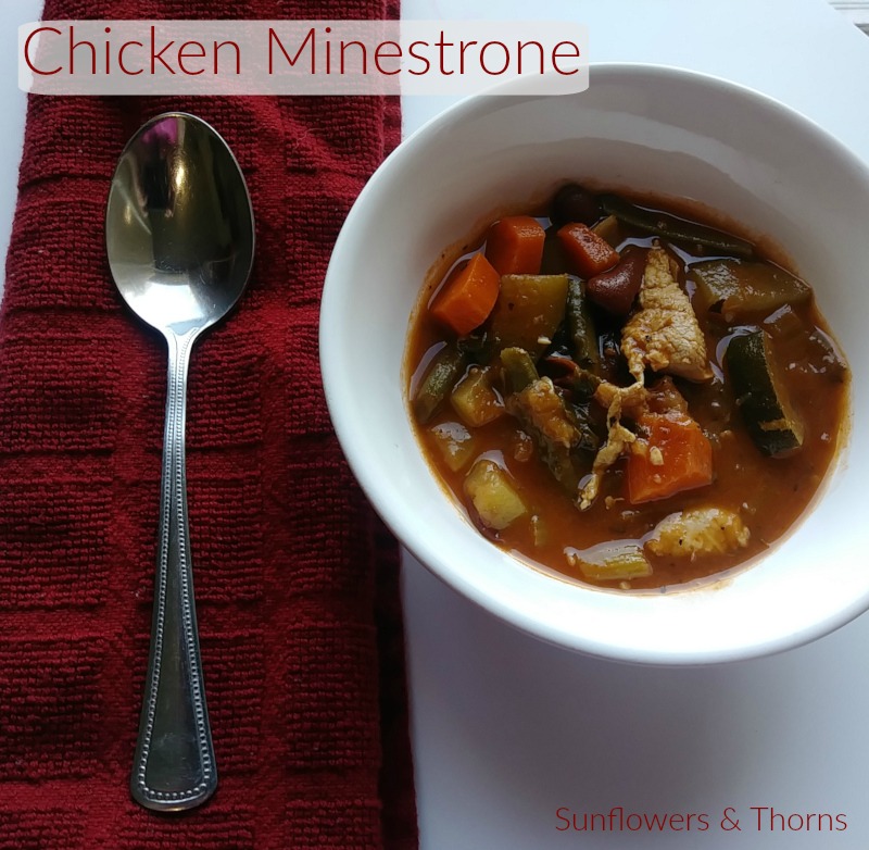 White bowl with chicken and vegetable minestrone in it. Bowl is next to a spoon on top of a red napkin