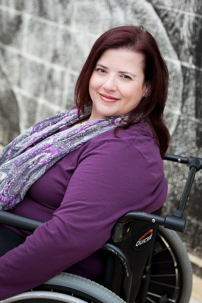Woman in a wheelchair smiling at the camera with confidence. She is wearing a purple shirt and a scarf. 